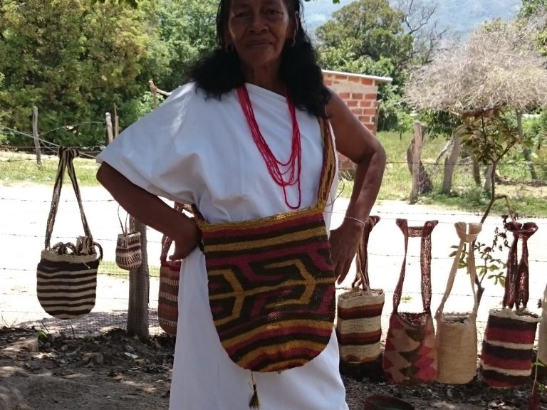Femme portant un sac traditionnel coloré, vêtue d'une robe blanche, avec des colliers.